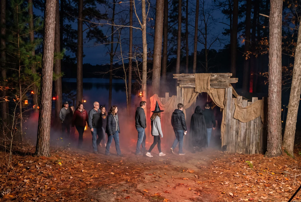 Costumed visitors walking a fog-filled haunted trail at night through tall pine woods, lit by red and orange light, with a lake glinting through the trees