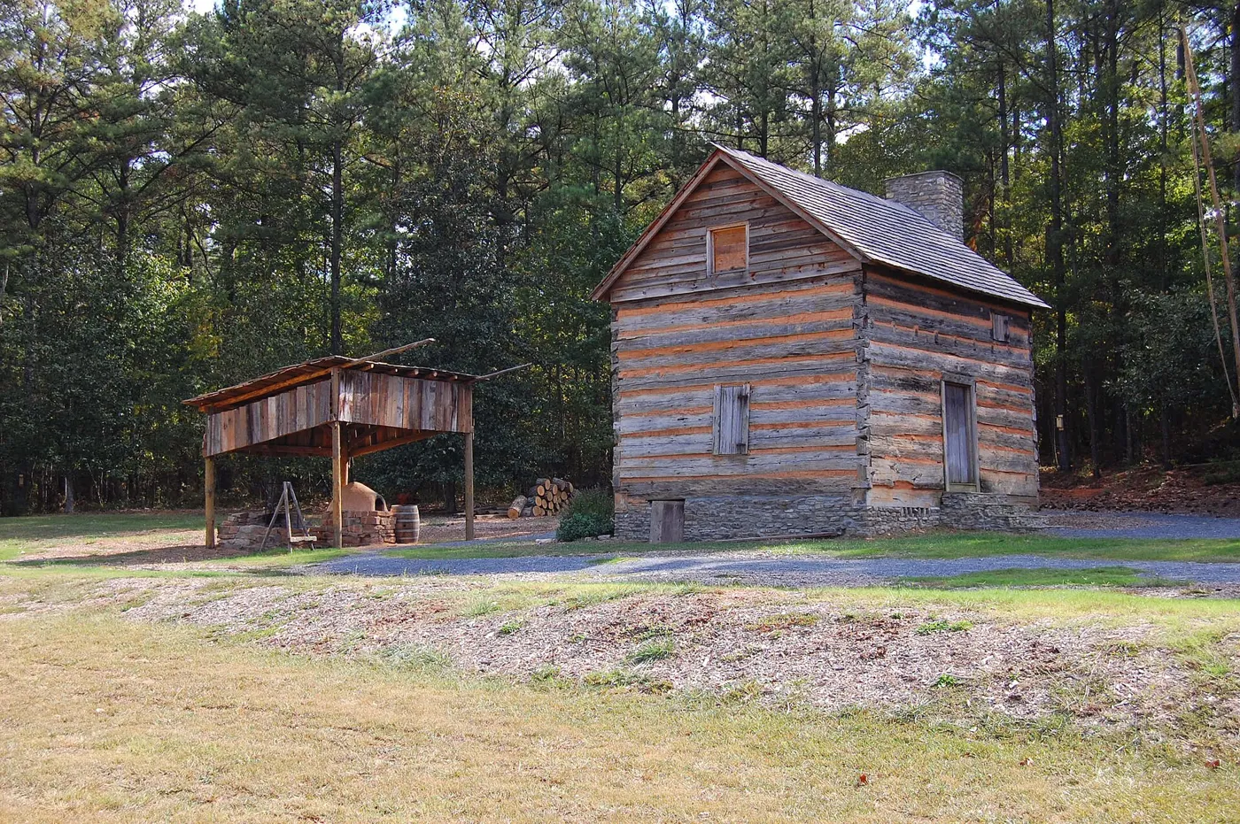 A two-story log blockhouse with a wood shingle roof standing in a grassy clearing among trees