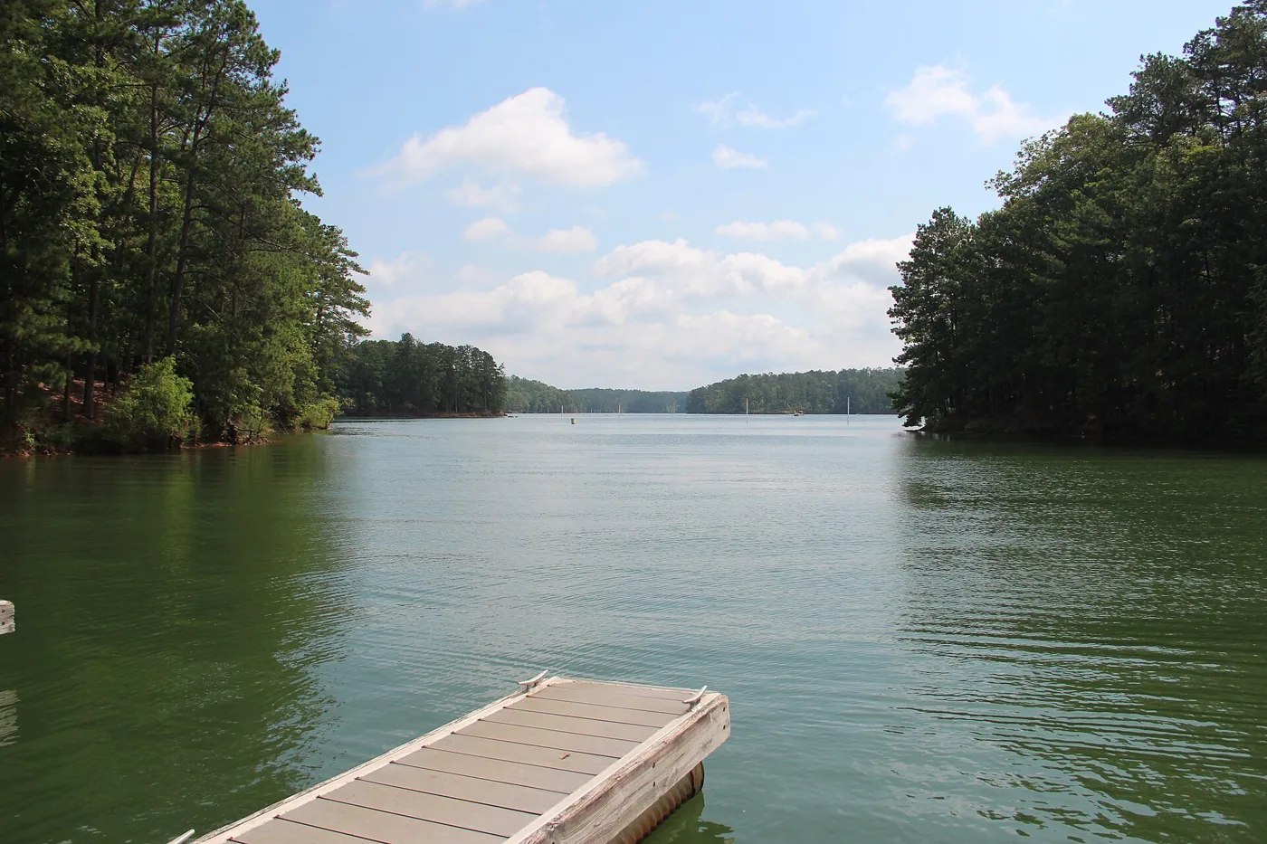 A floating dock in the foreground of a wide green lake ringed by forested hills under a blue sky