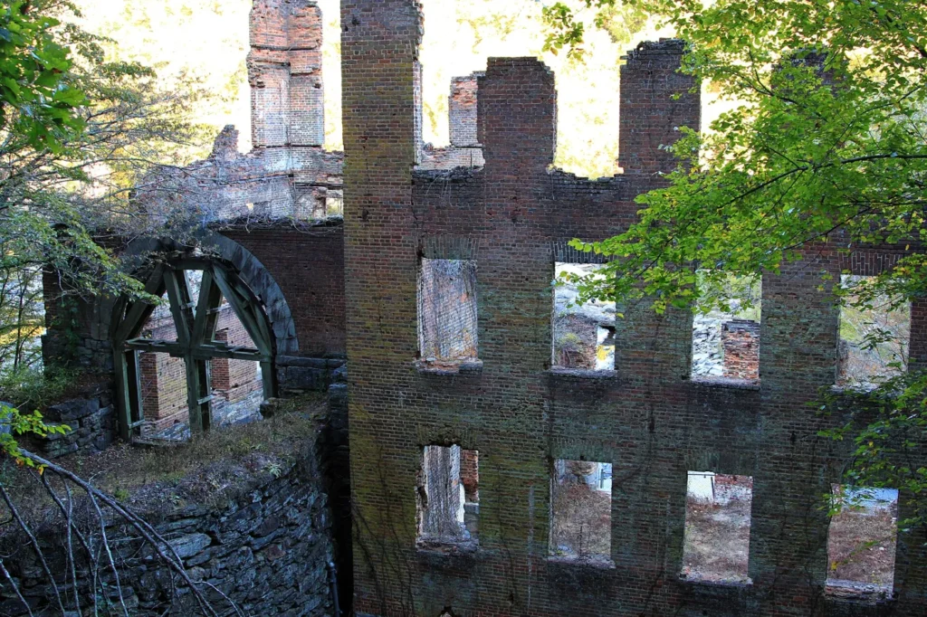 The roofless brick walls and empty windows of a burned textile mill beside a wooden water wheel, framed by green leaves